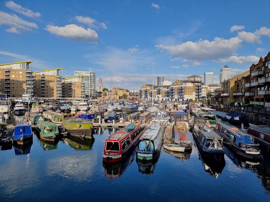 A scenic view of modern residential apartments with yellow brick facades and large glass balconies, situated along a river. Several boats are docked along the waterfront, including a blue and white ve