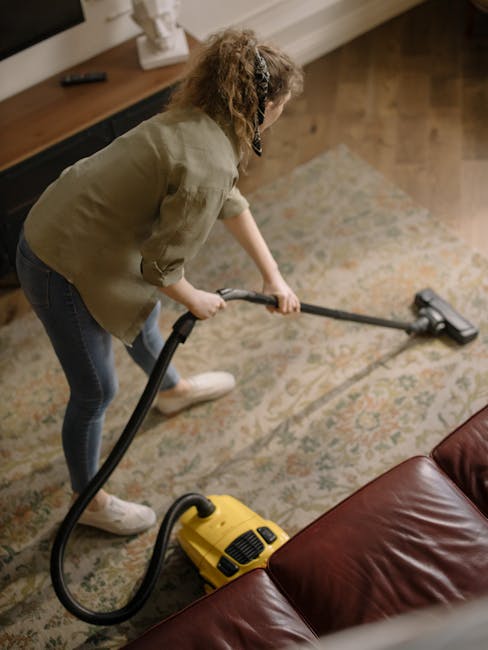 A woman operating a yellow vacuum cleaner on a traditional patterned area rug in a living room, with wooden flooring and a leather sofa visible to the side. The scene depicts surface cleaning and deep